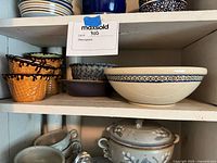 Shelf with three novelty ice cream waffle cone bowls with chocolate drip glaze on edges, a blue and white speckled bowl, a large cream bowl with geometric border pattern, a dark solid bowl and a blue speckled bowl.