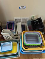 Photo showing stack of multiple plastic baskets in assorted colors and sizes arranged against a wall.