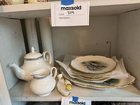 Photo of the white ceramic tea set components including teapot, creamer, strainer, and a stack of decorative plates on a shelf.