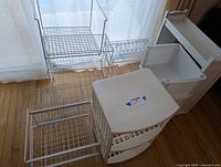Photo showing a set of white wire and plastic shelving units and components arranged on the floor near a window with natural light. Includes metal wire shelves, plastic units with drawers and open compartments.