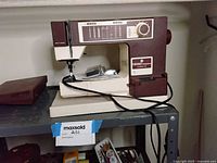 Viking Husqvarna Classica 100 sewing machine on shelf with corded power source, foot pedal on shelf next to it, plastic bag of accessories on machine bed