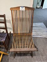 Wooden slat-back lounge chair with vertical slats for seat and backrest, showing slight recline and curvature at top of backrest, finished in dark wood stain, positioned on carpeted floor inside a room with adjacent furniture visible.