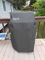 Closed Weber grill covered with black Weber-branded protective cover on outdoor deck.