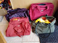 Four assorted women's travel bags arranged on the floor. Colors include deep purple, red, dark pink, and olive green. Different bag styles including totes and duffle bags.