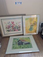 Three framed art prints arranged on floor: one floral garden scene, one flower bouquet in blue vase, and one rural barn landscape.