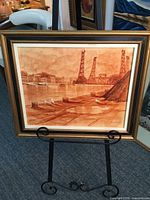Framed sepia-toned oil watercolour painting of a canal with industrial and waterfront buildings, birds, dock with posts, and cranes, displayed on a black decorative easel.