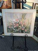 Photo depicting framed artwork by Nancy O’Toole on an easel, showing floral scene with pink and white flowers and green foliage.