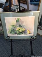 Framed watercolor painting of pear and grapes placed on a black metal easel, showing full front view with aluminum frame and pale green mat.