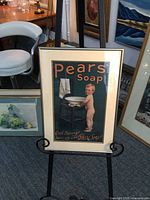 Photo showing the framed Pears Soap vintage advertising poster with a boy blowing bubbles, displayed on an easel with other framed art in the background.