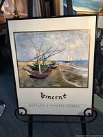 Photo of framed Van Gogh poster titled Bateaux A Saintes Maries showing colorful boats on the beach under a cloudy sky, in black frame with white and gold mat.