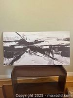 Abstract black and white acrylic painting on canvas displayed horizontally on a small wooden stand against a beige wall.