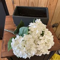Artificial white hydrangea flower bouquet with clusters of white petals and green leaves placed on a wooden surface showing the fullness and detail of the petals and leaves