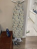 Full view of ironing board with polka dot fabric cover featuring circular patterns in various colors, white frame, iron rest, and Black+Decker iron with cord placed at base of board
