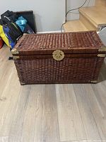 Full view of rectangular brown wicker trunk/chest with brass corner accents and brass latch on front, placed on light hardwood floor near stairway.
