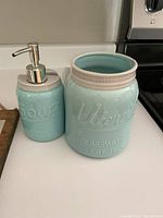 Side view of light blue ceramic soap dispenser and utensil holder with embossed text and off-white rim on counter.