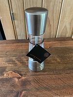 Front view of a stainless steel and acrylic Cole & Mason pepper grinder on a wooden surface showing the black brand tag.