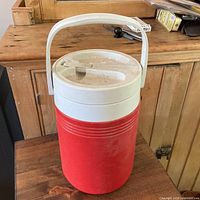 Front view of red and white insulated water jug cooler showing screw-on lid with flip-open spout and swing-up handle.