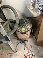 Full view of the Craftsman wet/dry shop vacuum showing the cylindrical red and black tank and the attached flexible grey hose next to a ladder and other items in a dusty workshop area.