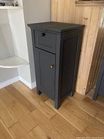 Full side-angle view of dark grey nightstand sitting on wooden floor, showing a drawer with black pull and cabinet door with brass knob.