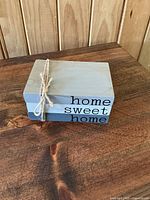 Image of a rectangular gray wood block sign resting on a wood table, showing the phrase 'home sweet home' in black lettering, tied with a natural fiber twine.