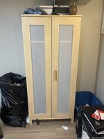 Closed IKEA wardrobe in light wood with frosted panels on two doors, standing on wooden floor, with surrounding household items visible.
