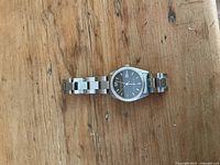 Kenneth Cole stainless steel wristwatch laying flat on wooden surface, showing front face and band spread out horizontally.