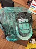 Front view of Keurig coffee maker wrapped in green plastic wrap, showing the control panel and brand logo.