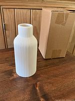 Single off-white ceramic vase with vertical ribbed texture standing on wooden surface next to a plain brown cardboard box.
