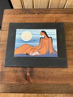 Photo of front of art card mounted on black mat board showing Indigenous woman and child with a white moon, on a rustic wood table.