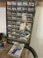 Front view of black plastic drawer organizer with 47 plastic drawers (39 small, 8 large) filled with assorted metal fasteners including screws, nuts, bolts, washers, some in small plastic bags, with a dusty surface. A blue and white hot glue gun in packaging is visible at base of organizer.