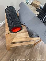 Foam roller and yoga mat displayed on wooden surface with floor visible in background showing foam roller black with red interior and rolled gray yoga mat.