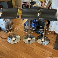 Three modern bar stools with black faux leather seats and chrome bases, arranged side by side on a wooden floor.