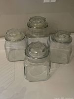 Four vintage clear glass storage jars with matching patterned glass lids arranged together on a surface.