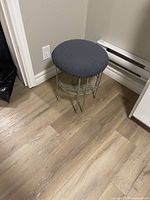 Full view of silver metal vanity stool with black padded fabric seat and integrated wire mesh shelf, placed on wooden floor near a wall outlet.
