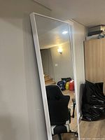 Full length vertical mirror with white frame leaning against the wall in a room showing the mirror surface and surroundings.