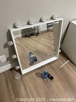 White framed square vanity mirror with large round bulbs around the frame, placed on floor showing both front and slight angled view.