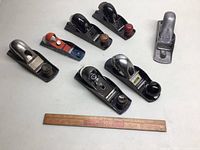 Top view of seven block planes arranged on a white background with a wooden ruler for scale.