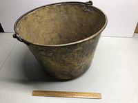 Front oblique view of the brass pail showing general shape and iron bail handle with ruler for scale.