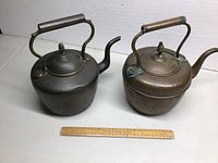 Two vintage brass kettles side by side on white surface, showing size comparison with a ruler in front.