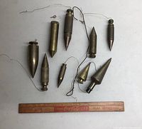 Nine brass plumb bobs on white surface with Coca-Cola ruler for scale, showing variety in sizes, shapes, and attached hanging wires.