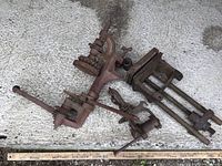 Wide shot showing all three vintage cast iron tools on a concrete surface: a canner, a bench vise, and a fence stretcher.