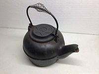 View of black vintage cast iron kettle with coiled metal handle and embossed lid.