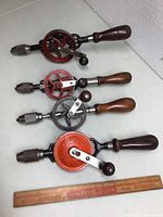 Four vintage eggbeater hand drills, each with wooden handles and metal gears, shown with a wooden ruler for scale.