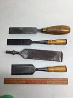 Four wood chisels laid flat with ruler for size reference, showing varying blade sizes and wooden handles.