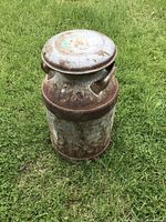 Front angled view of vintage metal milk can on grass, showing rust, lid and handles