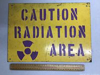 Front side of the metal sign with 'CAUTION RADIATION AREA' stencil and radiation symbol in purple on yellow background, ruler for scale.