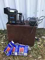Photo showing Polaroid Studio Express camera on top of brown carrying case with power supply and three unopened Polaroid 100 film packs in front.