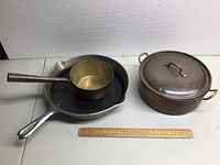 Cast iron pan with small brass pot inside and copper pot with lid next to it, ruler for scale.