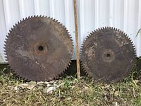 Two large buzz saw blades shown side by side outside against a white wall. Larger blade includes measuring stick showing 26-inch diameter.