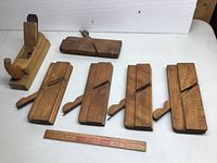 Six wooden planes arranged on a flat surface, including five flat sole wooden planes and one wooden plane with a curved handle and metal blade, with a vintage Coca-Cola ruler for scale.
